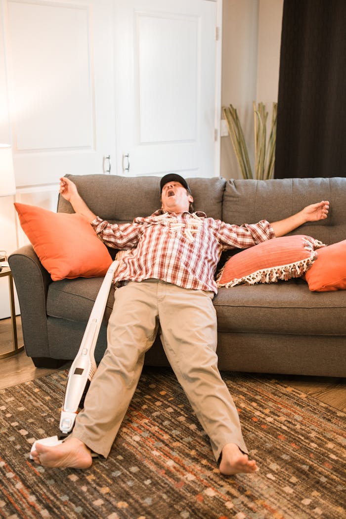 Tired adult man taking a nap on the sofa after cleaning, surrounded by orange pillows.